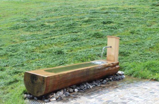 Holzbrunnen mit fließendem Wasser auf Steinpflaster vor frisch gemähter Wiese.