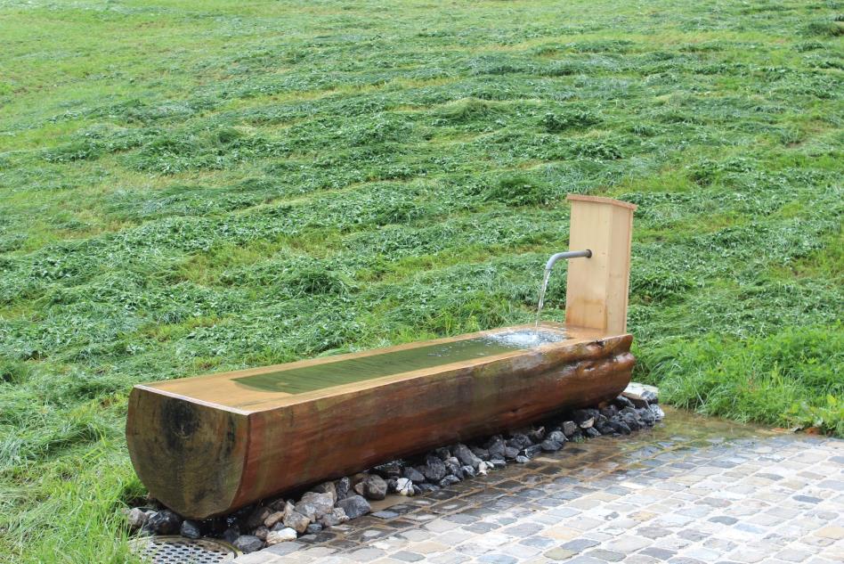 Holzbrunnen mit fließendem Wasser auf Steinpflaster vor frisch gemähter Wiese.