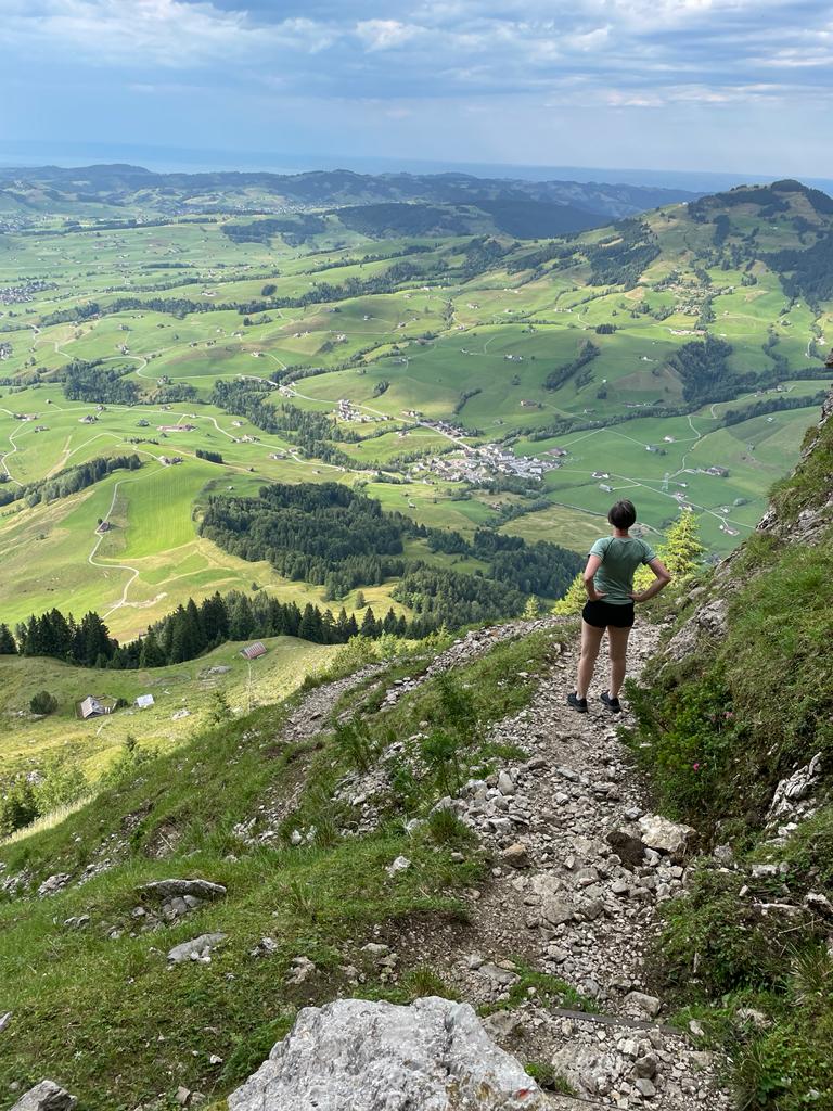 Wanderin steht am steinigen Pfad mit weitem Blick über das grüne Tal.