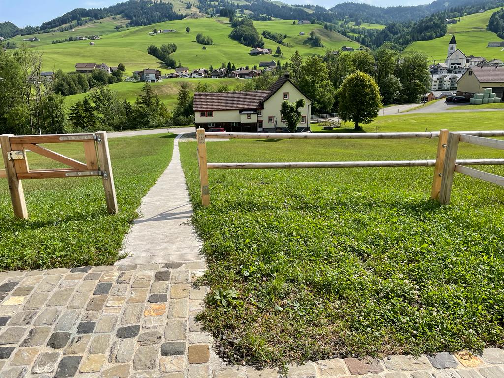 Blick über einen gepflasterten Weg mit Holzzaun auf ein Dorf in sanfter Hügellandschaft.