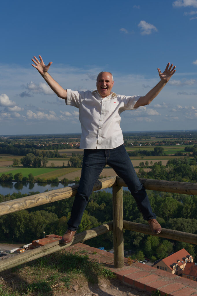 Mann steht freudig auf einem Geländer mit weitem Ausblick.