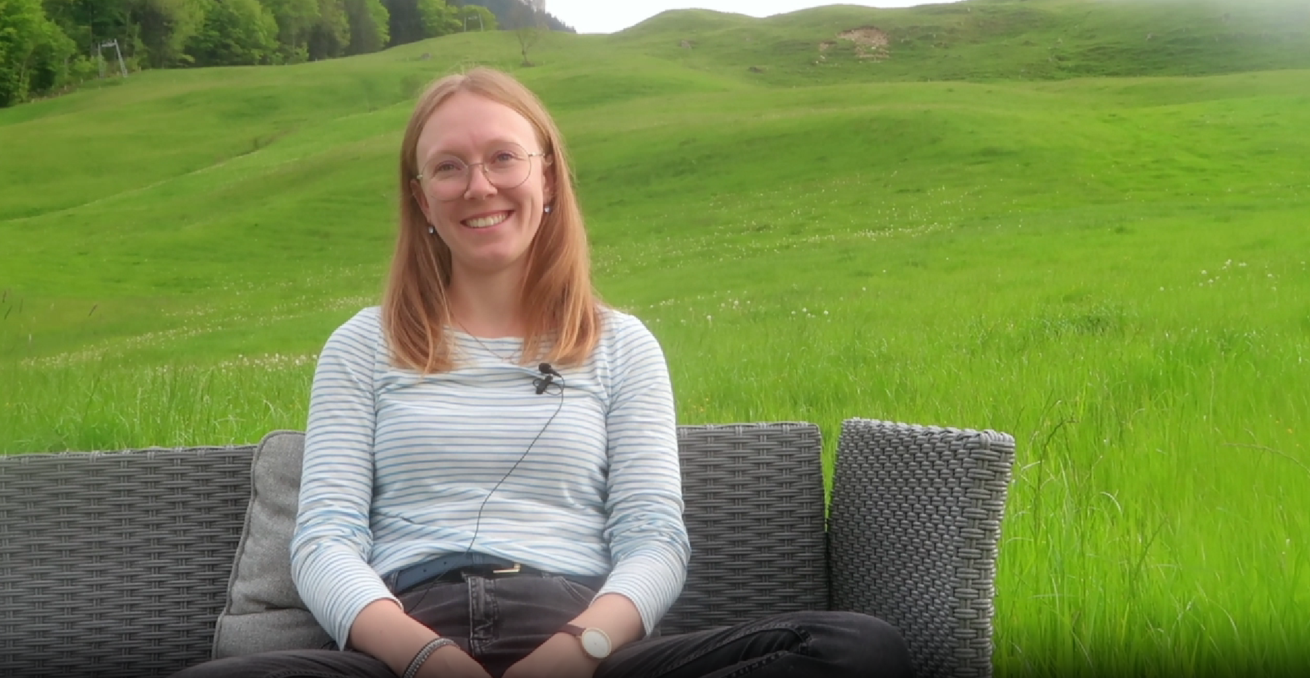 Junge Frau mit Brille lächelt auf Outdoor-Sofa vor sanfter Hügellandschaft.