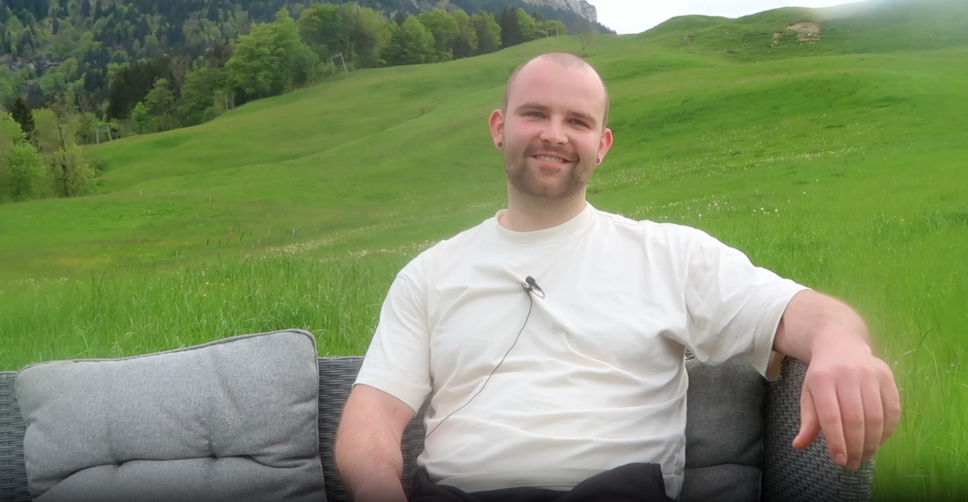 Mann im weißen Shirt sitzt entspannt auf Outdoor-Sofa vor grüner Berglandschaft.
