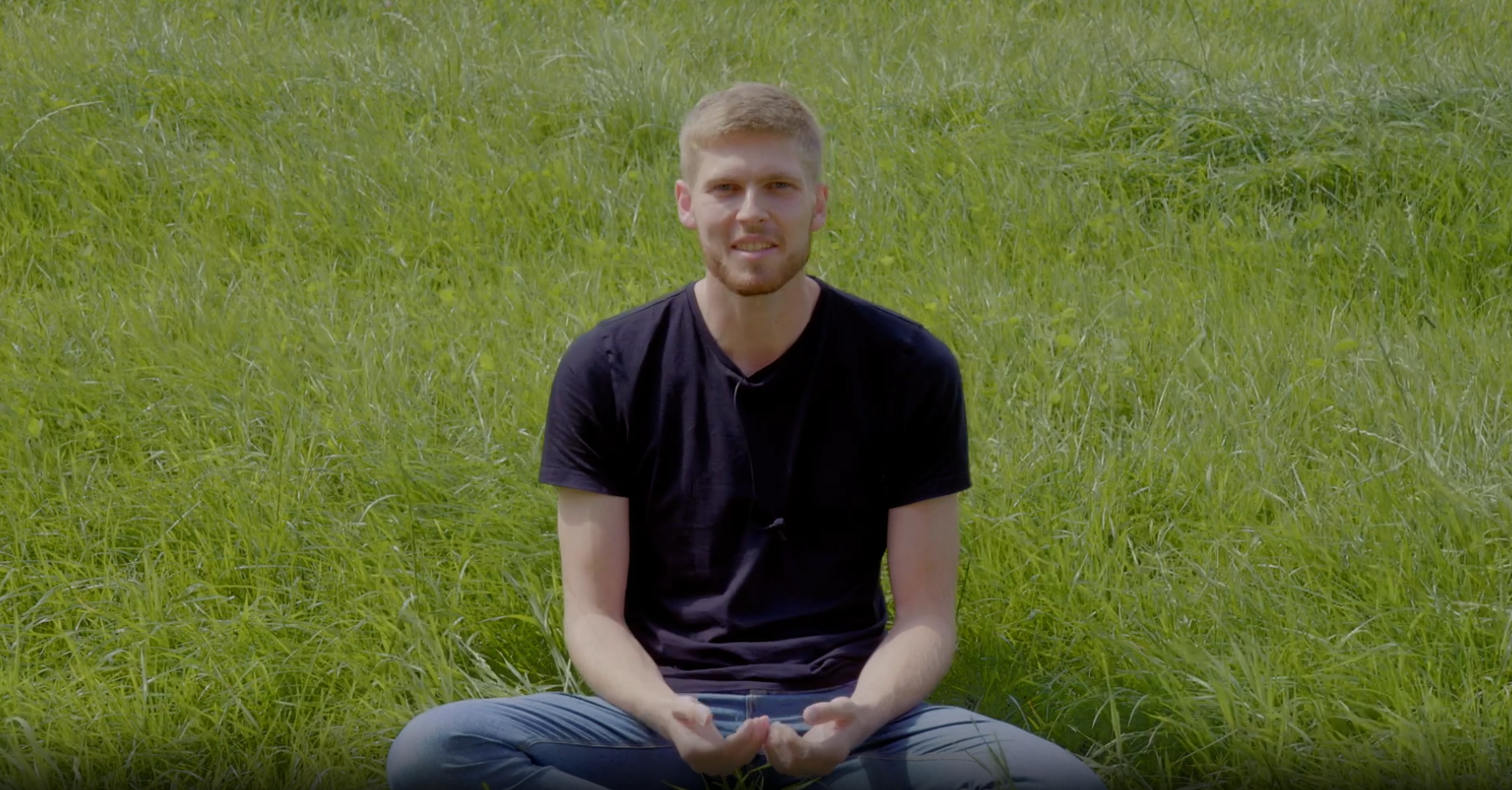 Junger Mann im schwarzen Shirt sitzt lächelnd im Gras und spricht zur Kamera.